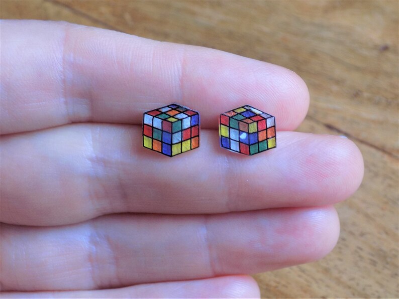 Puede incluir: Un par de pendientes de bot&oacute;n con un dise&ntilde;o de cubo de Rubik colorido. Los pendientes est&aacute;n hechos de pl&aacute;stico transparente con un dise&ntilde;o impreso.