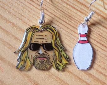 May include: A pair of earrings featuring a cartoon illustration of a man with long blonde hair wearing sunglasses and a bowling pin. The earrings are made of clear plastic with silver hooks.