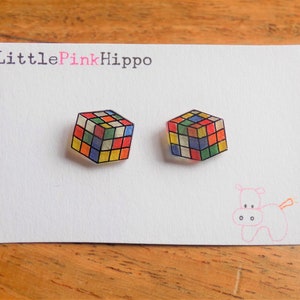 Puede incluir: Un par de pendientes con un dise&ntilde;o de cubo de Rubik colorido. Los pendientes est&aacute;n hechos de acr&iacute;lico transparente y tienen un fondo blanco. El texto "LittlePinkHippo" est&aacute; impreso en la tarjeta.