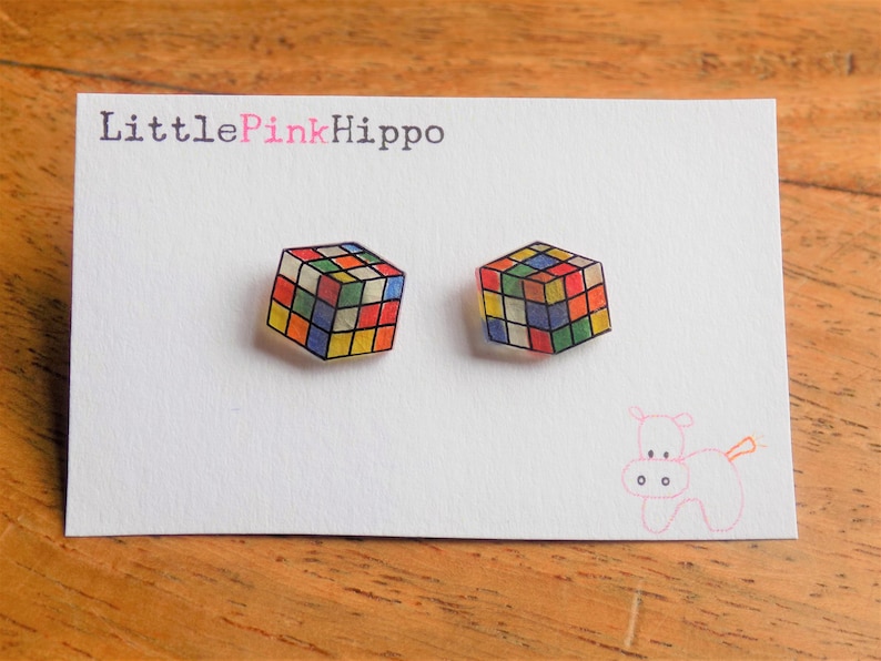 Puede incluir: Un par de pendientes con un dise&ntilde;o de cubo de Rubik colorido. Los pendientes est&aacute;n hechos de acr&iacute;lico transparente y tienen un fondo blanco. El texto "LittlePinkHippo" est&aacute; impreso en la tarjeta.