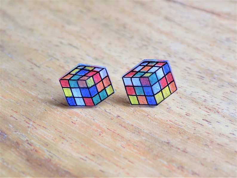 Puede incluir: Un par de pendientes de cubo transparente y colorido. Cada pendiente es una representaci&oacute;n 3D de un cubo de Rubik con una variedad de colores, incluyendo rojo, azul, amarillo, verde y naranja.