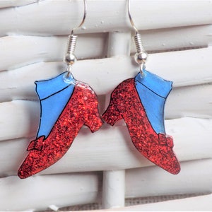 Op de afbeelding: Een paar rode glitteroorbellen in de vorm van robijnpantoffels met blauwe toppen. De oorbellen hebben zilveren haakjes.