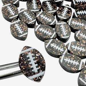 May include: A collection of small, football-shaped embellishments. Each piece is dark brown with gold glitter and white stripes, resembling a football. One is held by tweezers, showcasing the detail.