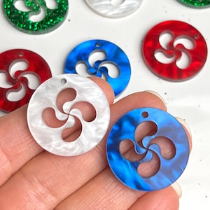 May include: A collection of colorful, round charms featuring a stylized Basque cross design. The charms are in various colors, including green glitter, red, white, and blue. Each charm has a small hole for stringing.