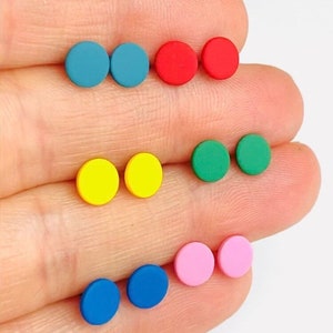 May include: A collection of colorful stud earrings in various shades, including blue, red, yellow, green, and pink. Each pair of earrings is a small, solid circle. The earrings are displayed on a light-colored background.