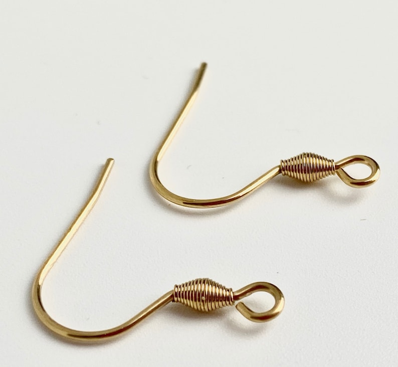 K&ouml;nnte beinhalten: Zwei goldfarbene Ohrhaken. Jeder Haken hat ein gebogenes Drahtdesign mit einem gewickelten Drahtdetail in der N&auml;he der Schlaufe. Die Haken sind f&uuml;r die Schmuckherstellung konzipiert und werden vor einem wei&szlig;en Hintergrund gezeigt.