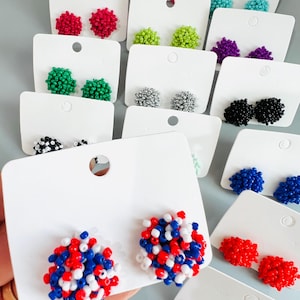 May include: A collection of colorful beaded stud earrings displayed on white cards. The earrings come in various colors, including red, blue, white, green, and black. The earrings are round and clustered, resembling small pom-poms.