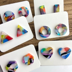 May include: A collection of colorful resin earrings in various shapes, including circles, rectangles, triangles, and hearts. Each earring features a unique pattern of mixed colors, mounted on white cards.