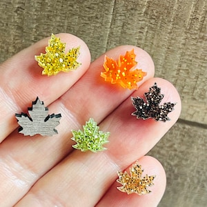 May include: A collection of small, maple leaf-shaped embellishments in various colors and textures. The leaves include glittery gold, orange, and brown, as well as a wooden-look gray leaf and a green glitter leaf. These are likely for crafting or jewelry making.