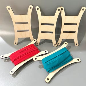 May include: Wooden thread spool holders with red and teal elastic bands. The holders are made of light-colored wood and have a unique shape with slots and holes. The elastic bands are neatly wound on the holders, ready for use.