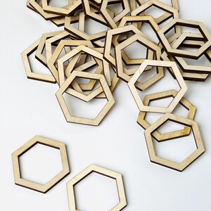 May include: A collection of light-colored wooden hexagon frames scattered across the frame. Each piece is a thin, flat, open-centered geometric shape with smooth edges and a natural wood grain finish, showing a consistent light tan color throughout.