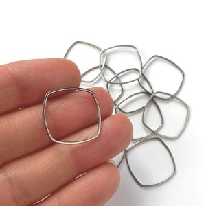 May include: A collection of silver-colored, square-shaped metal jewelry findings. The findings have rounded corners and are displayed against a white background. One finding is held in a hand.