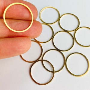 May include: A collection of small, gold-colored metal circle charms. The charms are thin and have a smooth, polished finish. The circles are arranged on a white surface, with one held by a hand.