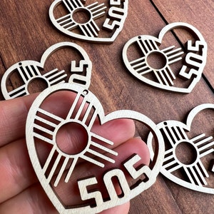 May include: Wooden heart-shaped ornaments with a cut-out design featuring a sun symbol and the number "505". The ornaments are made of light-colored wood and have a small hole for hanging. The design is a symbol of New Mexico.