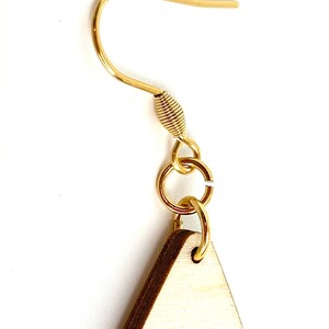 K&ouml;nnte beinhalten: Nahaufnahme eines goldfarbenen Ohrringhakens mit einem kleinen, daran befestigten Holz-Dreieck. Der Ohrringhaken besteht aus einem d&uuml;nnen, gebogenen Draht mit einem gewickelten Abschnitt in der N&auml;he der Oberseite. Das Holz-Dreieck hat einen hellbraunen Rand.