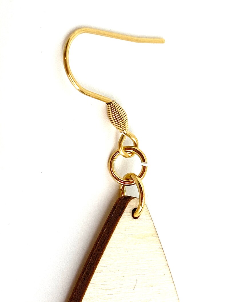 K&ouml;nnte beinhalten: Nahaufnahme eines goldfarbenen Ohrringhakens mit einem kleinen, daran befestigten Holz-Dreieck. Der Ohrringhaken besteht aus einem d&uuml;nnen, gebogenen Draht mit einem gewickelten Abschnitt in der N&auml;he der Oberseite. Das Holz-Dreieck hat einen hellbraunen Rand.