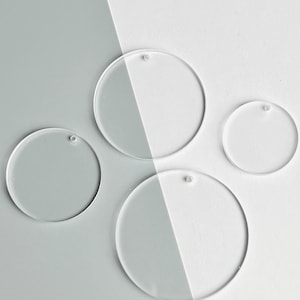 May include: Four clear acrylic circle blanks with small holes for hanging. The circles vary in size and are arranged on a two-tone background of gray and white. These blank circles are ideal for crafting and DIY projects.