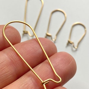 May include: Close-up of several gold-colored earring hooks in varying sizes. The hooks are made of thin wire, with a curved top and a small, straight post at the bottom. The hooks are held in a hand against a white background.