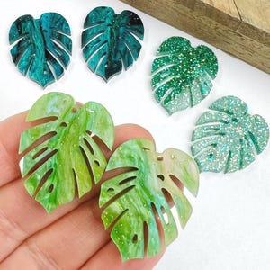 May include: A collection of six monstera leaf-shaped charms in various finishes. Two are dark teal with a marbled effect, two are bright green with a marbled effect, and two are translucent with green and silver glitter embedded throughout.