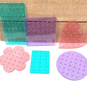 May include: A collection of colorful, glittery acrylic shapes, including a rectangle, a heart, a flower, a square, and a circle. The shapes are in shades of green, purple, red, pink, and turquoise. Each shape has small, evenly spaced holes.