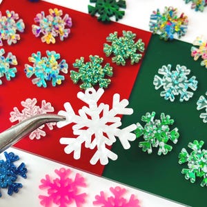 May include: A collection of small, flat snowflake-shaped charms in various colors including blue, green, pink, white, and iridescent finishes. A pair of metal tweezers holds a single white snowflake charm, which features a small hole at the top.