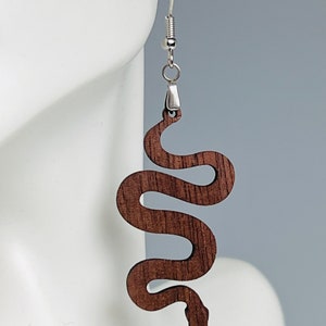 May include: A single wooden earring in the shape of a snake. The earring is a dark brown color and hangs from a silver-colored hook. The snake design is detailed with visible wood grain.