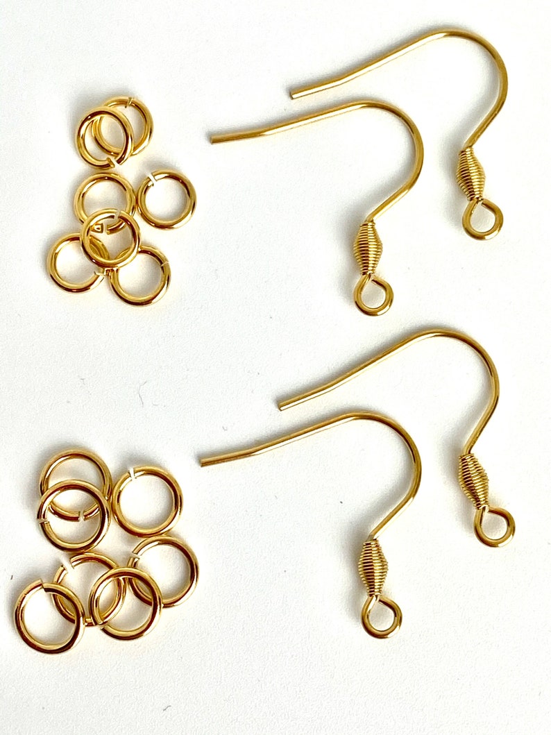 K&ouml;nnte beinhalten: Eine Sammlung goldfarbener Schmuckherstellungsutensilien, darunter mehrere offene Sprungringe und Ohrhaken. Die Ohrhaken haben ein gebogenes Design mit einem spiralf&ouml;rmigen Detail am unteren Ende. Die Sprungringe sind klein und rund.