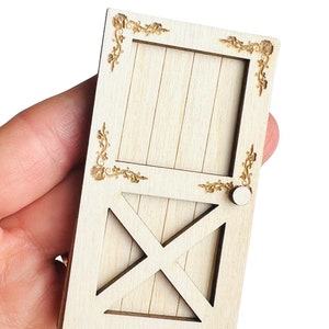 May include: A miniature wooden door with a light wood finish. The door features a decorative floral design in the corners and a cross-shaped accent. A small, round doorknob is visible. This item is likely for a dollhouse or craft project.