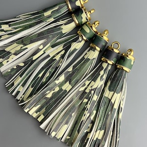 May include: Five camouflage print tassels with gold-colored hardware. The tassels feature a mix of green, black, and tan colors. The top of each tassel is wrapped in a matching camouflage pattern with a gold-colored ring.