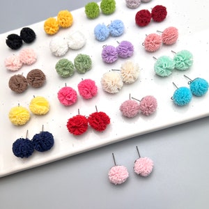 May include: A collection of colorful pom-pom stud earrings in various colors, including black, yellow, white, green, blue, purple, pink, red, and brown. The earrings are displayed on a white rectangular tray against a gray background. Each earring has a post.