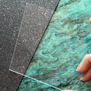 May include: A clear, rectangular acrylic sheet is held by a person's fingers. The sheet is positioned over a dark gray, speckled surface and a teal, marbled surface. The acrylic sheet is thin and transparent.