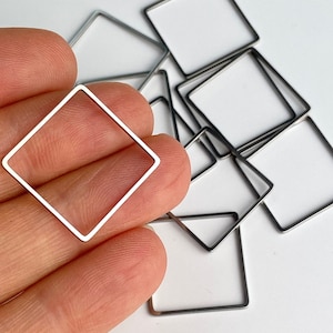 May include: A collection of silver-colored, square-shaped metal frames. One frame is held in a hand, with several others scattered in the background. The frames have a minimalist design, with clean lines and a geometric shape. The frames are likely for jewelry making or crafting.