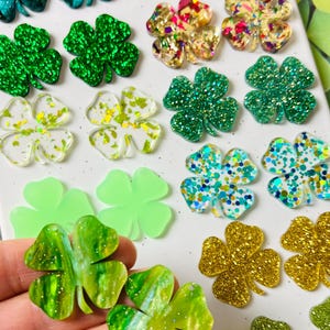 May include: A collection of various four-leaf clover designs in shades of green, gold, and iridescent colors. Some clovers have glitter or confetti accents, while others are translucent or solid. The clovers are arranged on a white surface.