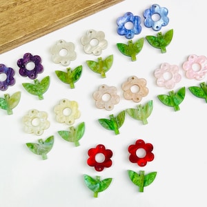 May include: A collection of colorful flower and leaf-shaped acrylic charms. The charms are in various colors, including purple, blue, pink, red, and green. The flowers have a hole in the center, and the leaves have a detailed vein pattern.