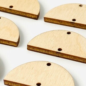 May include: Close-up of several unfinished wooden half-circle shapes with a hole drilled near the top. The wood has a light, natural color with a darker brown edge. These craft supplies are ideal for jewelry making or other DIY projects.