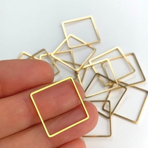 May include: A collection of small, gold-colored square metal frames. One frame is held in a hand, with several others scattered in the background. The frames are thin and have a minimalist design, suitable for jewelry making or crafting.