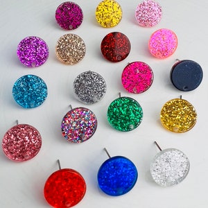 May include: A collection of colorful, round stud earrings with glitter embedded in clear resin. The earrings are in various colors, including pink, blue, green, and gold. Each earring has a silver post.