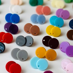 May include: A collection of colorful, round stud earrings in various shades, including red, blue, yellow, and purple. Each earring has a small hole near the top. The earrings are scattered across a white surface, creating a vibrant display.