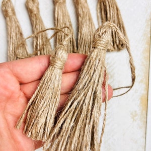 May include: Close-up of several handmade jute tassels in varying sizes. The tassels are a natural tan color, with tightly wound tops and long, fringed bottoms. They are arranged on a white surface, suggesting a craft or decor item.