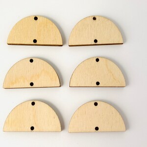 May include: Six unfinished wooden half-circle shapes with two small holes and one larger hole for hanging. The wood has a light, natural color and visible grain. These craft supplies are ideal for DIY projects.
