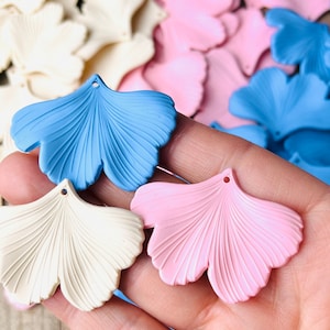 May include: Close-up of several Ginkgo leaf-shaped charms in various pastel colors: light blue, pink, and cream. Each charm has a small hole at the top for stringing. Other charms are visible in the background.