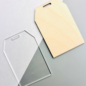 May include: Two blank tag-shaped objects. One is clear acrylic, the other is light-colored wood. Both have a small hole at the top for hanging. The objects are on a gray and white background.