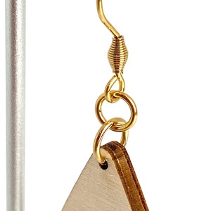 K&ouml;nnte beinhalten: Nahaufnahme eines goldfarbenen Ohrringhakens mit Spiraldesign, verbunden mit einem dreieckigen Ohrring aus Holz. Der Ohrring h&auml;ngt an einem silbernen Metallst&auml;nder. Das Holz hat eine helle, nat&uuml;rliche Farbe.