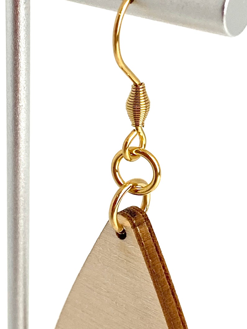 K&ouml;nnte beinhalten: Nahaufnahme eines goldfarbenen Ohrringhakens mit Spiraldesign, verbunden mit einem dreieckigen Ohrring aus Holz. Der Ohrring h&auml;ngt an einem silbernen Metallst&auml;nder. Das Holz hat eine helle, nat&uuml;rliche Farbe.