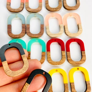 May include: A collection of oval-shaped pendants with a wood grain bottom and a colorful top. The pendants are in various colors, including orange, white, blue, pink, green, red, black, and yellow. Each pendant has a small hole at the top for stringing.