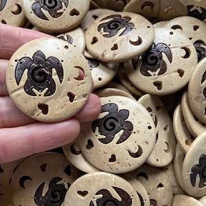 May include: Round, light-colored wooden beads with a turtle design carved in the center. The turtle is dark brown, and the beads have heart and crescent moon cutouts. A hand holds one bead, with many more in the background.