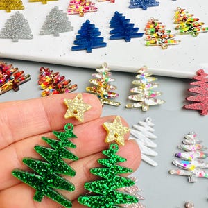 May include: A collection of Christmas tree-shaped charms in various colors, including green, pink, gold, and blue, with glitter accents. Some charms are solid, while others have a patterned design. A hand holds two green glittery tree charms with gold star toppers.