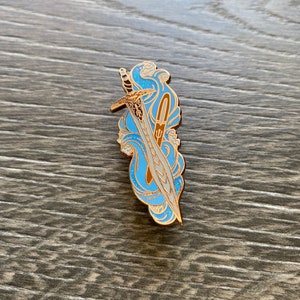 Op de afbeelding: Een goudkleurige metalen pin met een zwaardontwerp en een lichtblauw en wit golfpatroon. De pin heeft een gedetailleerd ontwerp met een gouden omtrek, tegen een houten achtergrond.