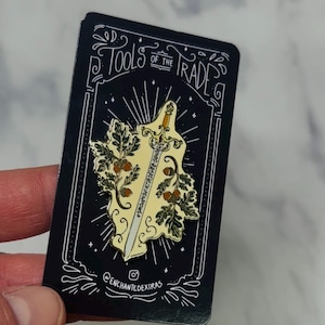 May include: A black rectangular pin with a sword design. The sword has a light yellow blade and a gold hilt, surrounded by oak leaves and acorns. The text reads "TOOLS OF THE TRADE" at the top and "@enchantedextras" at the bottom.