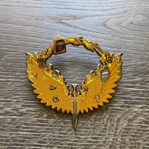 May include: A gold-coloured metal pin with a winged design. The wings are outlined in black and have a yellow flame pattern. A sword is in the centre of the wings. The wings are surrounding a book, a mirror, and other symbols.
